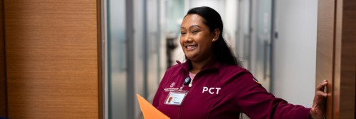 A patient care technician in a maroon uniform opens a door and greets a patient in a well-lit hallway.